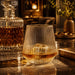Velora Tumbler Gift Set by The Line Edit. - A drink glass with a ridged texture, filled with amber liquid, sits on a marble surface beside a crystal decanter.
