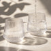Velora Tumbler by The Line Edit. - A drink bottle featuring a ribbed design, made of clear glass, with water being poured into two glasses.