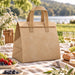 Wayfarer Cooler Bag - A brown cooler bag with a top handle, featuring a simple design. It has a logo and is set in a picnic environment.
