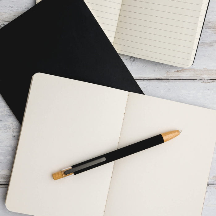 Serenity Aluminium Pen - A black aluminium pen rests on an open cream notebook, with a black notepad also visible in the background.