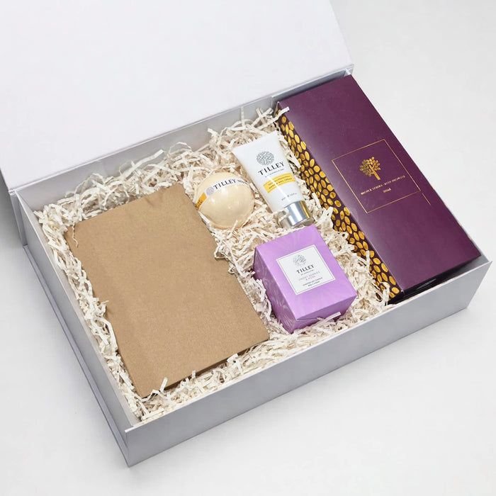The Mindful Moment - A gift box containing a brown notepad, a round item, a cream tube, a purple candle, and an item with a logo.