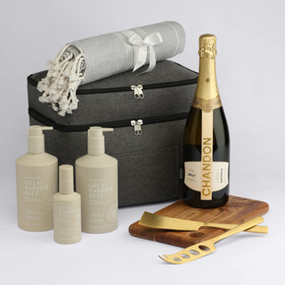 The Coastal Edit - A grey storage bag contains a throw blanket, champagne bottle, and skincare products in elegant packaging on a wooden tray with serving tools.