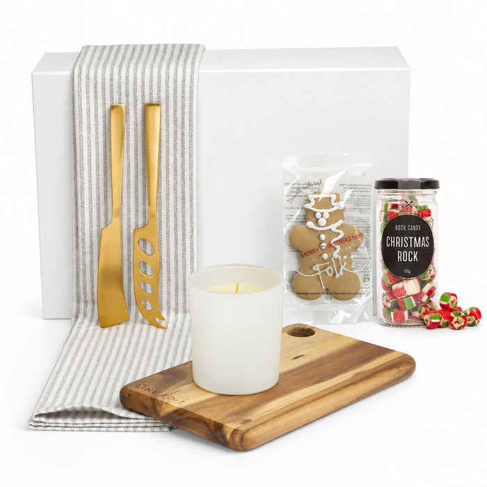 The Glow & Graze: Christmas Edition - A white box contains a wooden serving board, gold utensils, a candle, gingerbread, and Christmas sweets, all on a striped cloth.