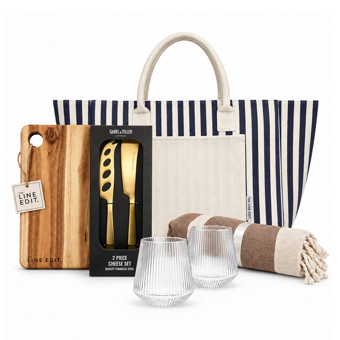 The Weekend Edit - A striped tote bag contains a cheese set, a wooden cutting board, two glass tumblers, and a striped blanket.
