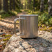 Bush Imitation Titanium Cup - A titanium cup rests on a rock, with a silver metallic finish, surrounded by greenery and a forest path.