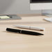 Axel Matte - A black pen and a white pen laid on a wooden desk, with a computer in the background. The pens have sleek designs.