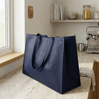 Jumbo Non Woven Shopper - A large navy blue non-woven shopper bag with sturdy handles rests on a kitchen counter. It features a subtle branding logo.