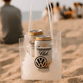 Icy Bags - Large - A clear cooler bag filled with ice and two cans, featuring a logo. Set on a sandy beach with people in the background.