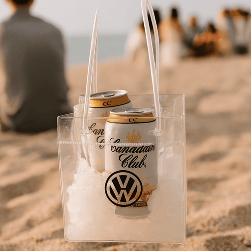 Icy Bags - Large - A clear cooler bag filled with ice and two cans, featuring a logo. Set on a sandy beach with people in the background.