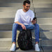 Swissdigital Vector Backpack - A man sits on steps, holding a phone, with a black Swissdigital Vector Backpack beside him.