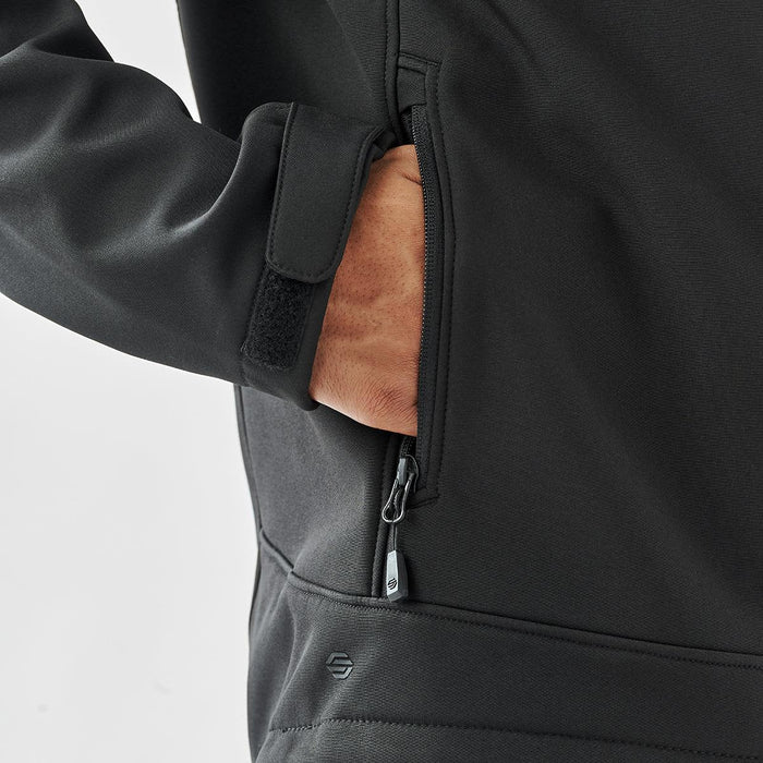 Men's Aleutian Softshell Jacket - A black softshell jacket with a zippered pocket, featuring a hand reaching inside the pocket.