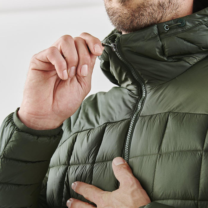 Men's Tundra Thermal Jacket - A close-up of a man adjusting the zipper on a dark green puffer jacket. The jacket features a high collar and quilted design.