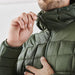 Men's Tundra Thermal Jacket - A close-up of a man adjusting the zipper on a dark green puffer jacket. The jacket features a high collar and quilted design.