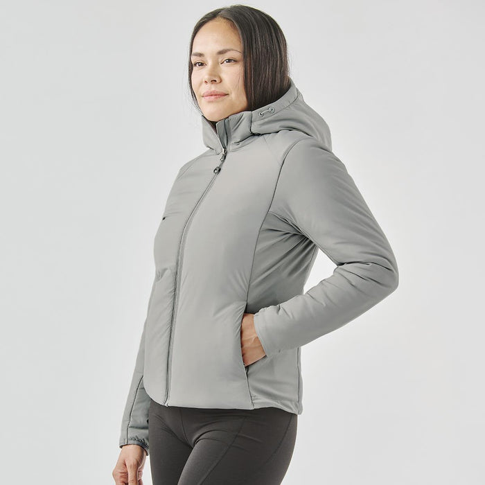 Women's Cascadia Thermal Jacket - A woman wearing a grey thermal puffer jacket with a hood, featuring side pockets and a fitted design.