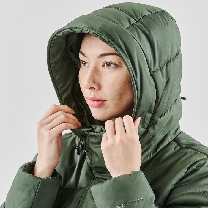 Women's Elkridge Parka - A woman wearing a green puffer jacket with a hood, adjusting it around her face. The jacket has a subtle logo.