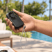 Mini Pocket Fan - A handheld black mini pocket fan with a wrist strap, being held outdoors near a pool.